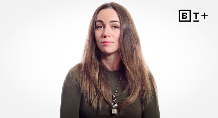 A woman with long brown hair wearing a dark top and necklace sits against a plain white background with a "B T +" logo in the top right corner.