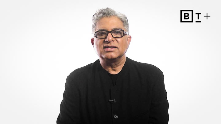 A person with short gray hair and glasses, wearing a black jacket, sits in front of a plain white background with a "B T +" logo in the top right corner.