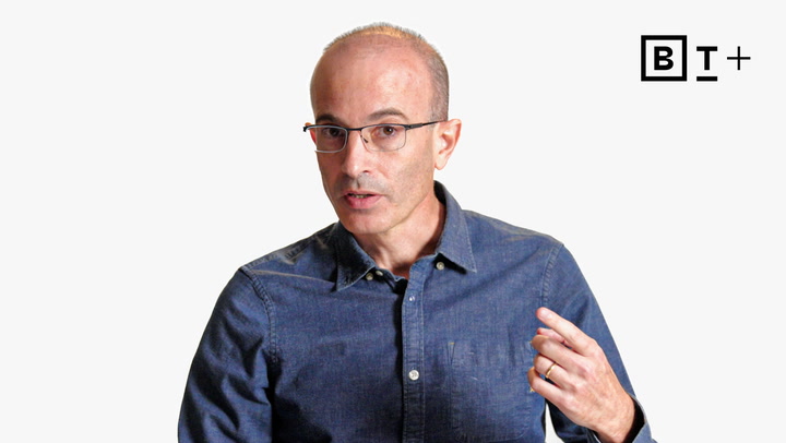 A man with glasses and a receding hairline wears a blue button-up shirt and gestures with his hand while speaking; a "B T +" logo appears in the upper right corner.