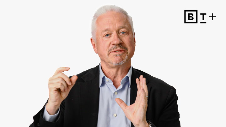 A man with short white hair and a beard, wearing a suit jacket and light blue shirt, gestures with his hands against a plain white background with a "B T +" logo in the corner.