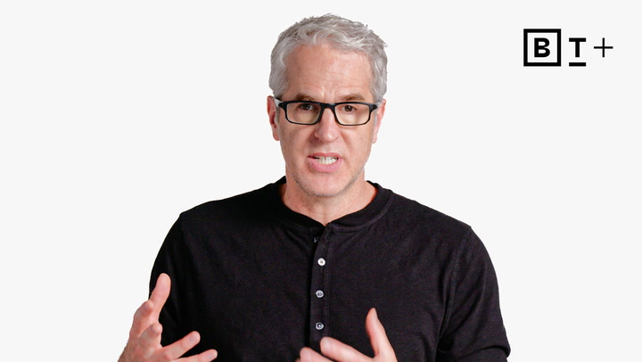 A man with gray hair and glasses, wearing a black henley shirt, speaks while gesturing with his hands; a "B T +" logo appears in the top right corner.