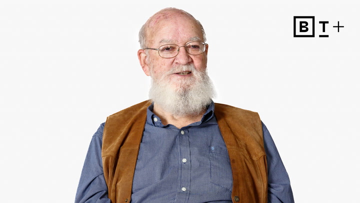 An older man with a white beard, wearing glasses, a blue shirt, and a brown vest, sits against a plain white background with a “B T+” logo in the corner.