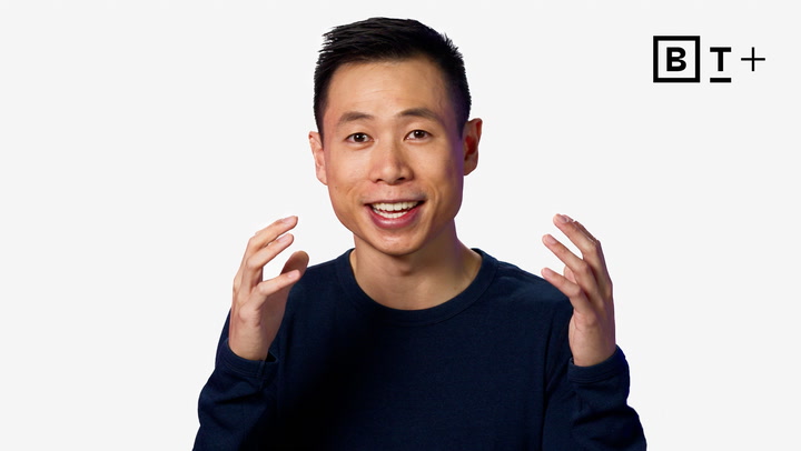 A person in a navy shirt gestures with raised hands while speaking in front of a plain white background with the "B T +" logo in the top right corner.
