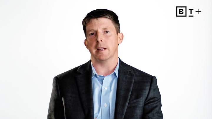 A man in a dark suit and blue shirt sits against a plain white background with a "BT+" logo in the top right corner.