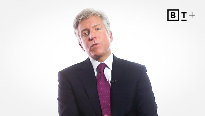 A man in a suit and tie sits against a plain white background with a "BT+" logo in the top right corner.