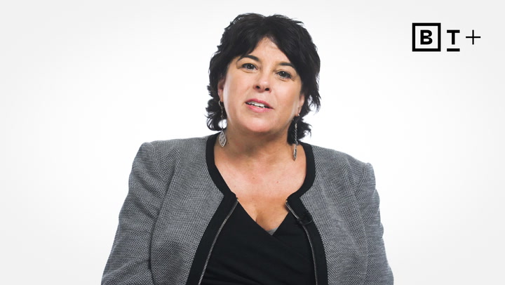 A woman with short dark hair wearing a grey blazer and black top sits against a plain white background with a "BT+" logo in the top right corner.