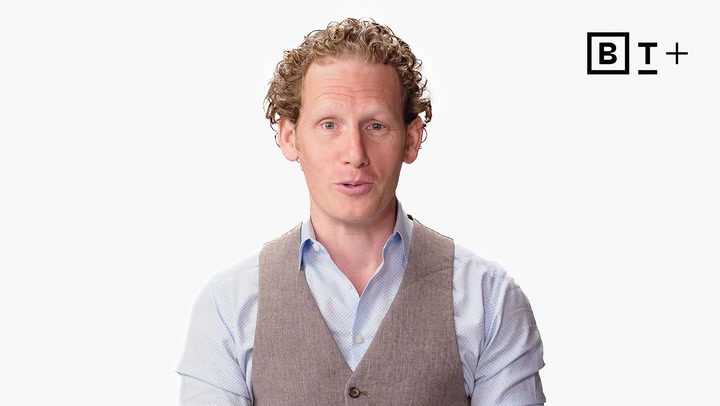 A man with curly hair wearing a light blue shirt and beige vest speaks in front of a plain white background with a "BT+" logo in the top right corner.