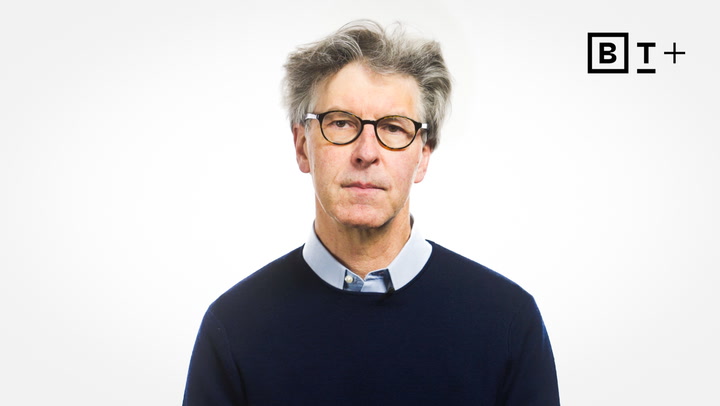 An older man with gray hair and glasses wearing a navy sweater over a collared shirt stands against a plain white background with a "BT+" logo in the top right corner.