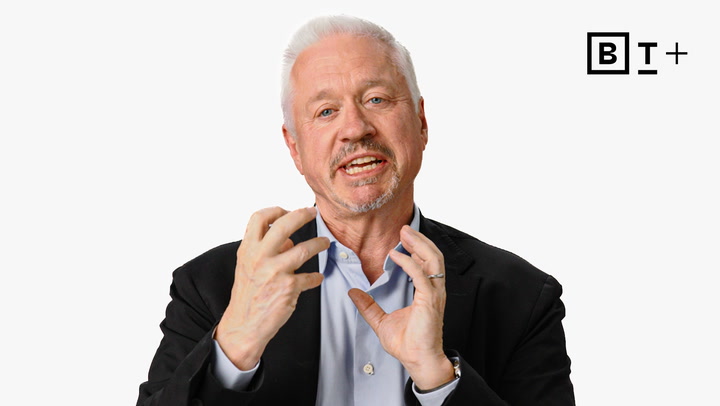 A man with short white hair and a trimmed beard, wearing a black blazer and blue shirt, gestures with both hands while speaking. "BT+" logo appears in the top right corner.