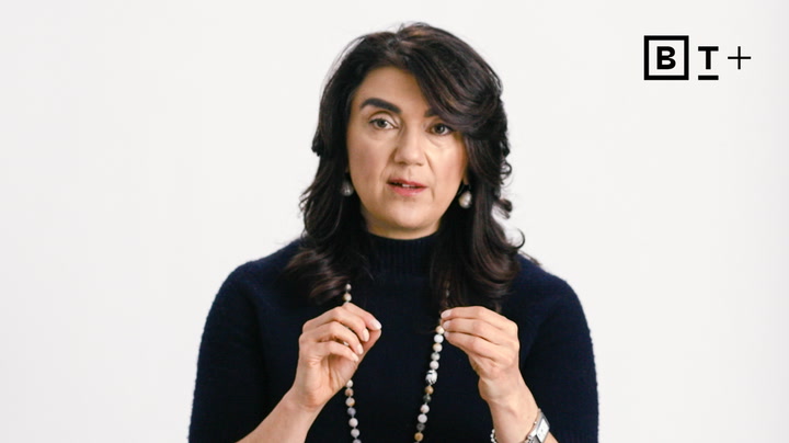 A woman with dark hair wearing a navy sweater and beaded necklace speaks while gesturing with her hands against a plain white background. The letters "B T +" appear in the top right corner.