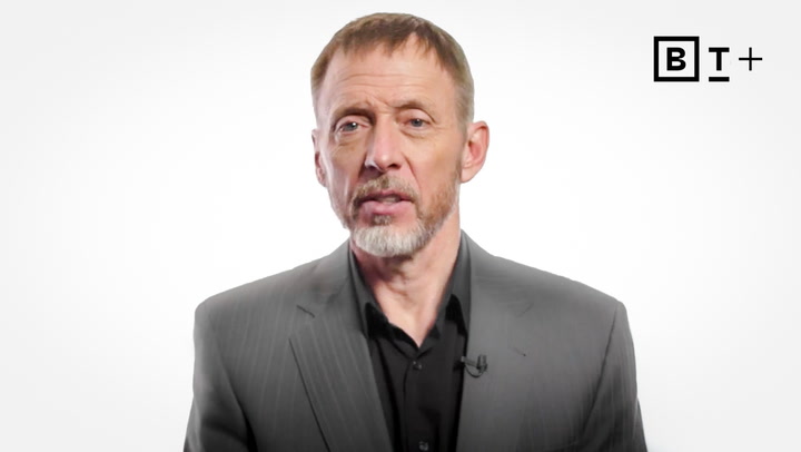 A man with short hair and a beard, wearing a gray suit jacket and black shirt, stands against a plain white background with a "BT+" logo in the top right corner.