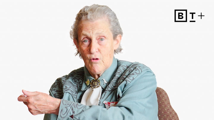 An older woman with gray hair wearing a patterned blue shirt and bolo tie sits in a chair, gesturing with her right hand. A "BT+" logo is visible in the top right corner.