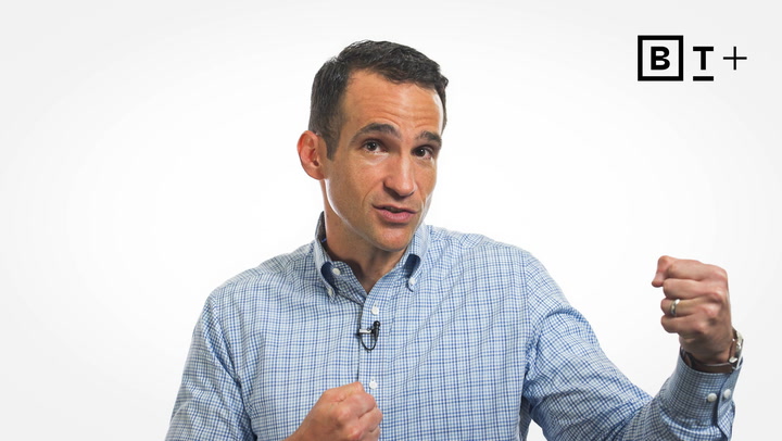A man in a checkered shirt gestures with his fists raised, standing against a plain white background with a "B T+" logo in the top right corner.