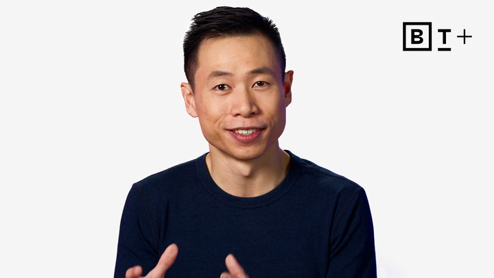 A man in a dark shirt speaks while gesturing with his hands. A black and white "BT+" logo is visible in the top right corner on a plain white background.