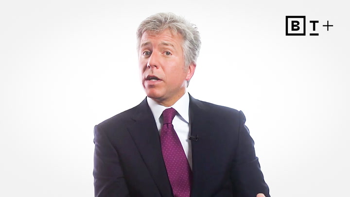 A man in a dark suit and purple tie sits in front of a white background with a BT+ logo in the top right corner.