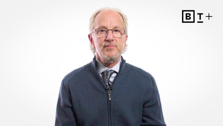 An older man with glasses and a trimmed beard, wearing a blue zip-up sweater and tie, sits in front of a plain white background with a "B T +" logo in the top right corner.