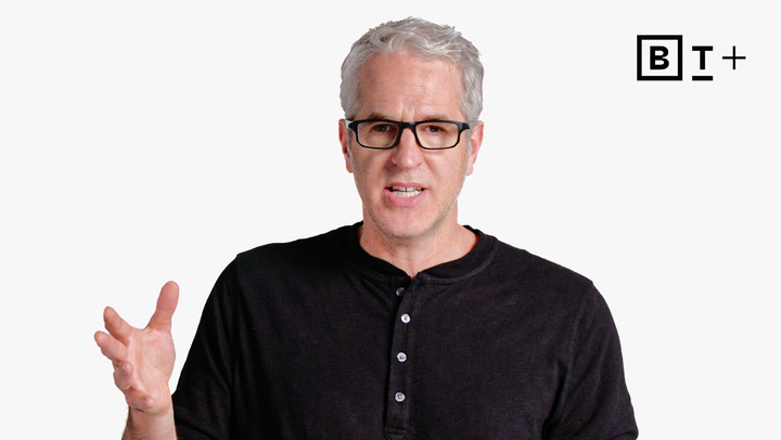A man with gray hair and glasses wearing a black henley shirt gestures with his left hand; a “B T+” logo appears in the upper right corner on a plain white background.