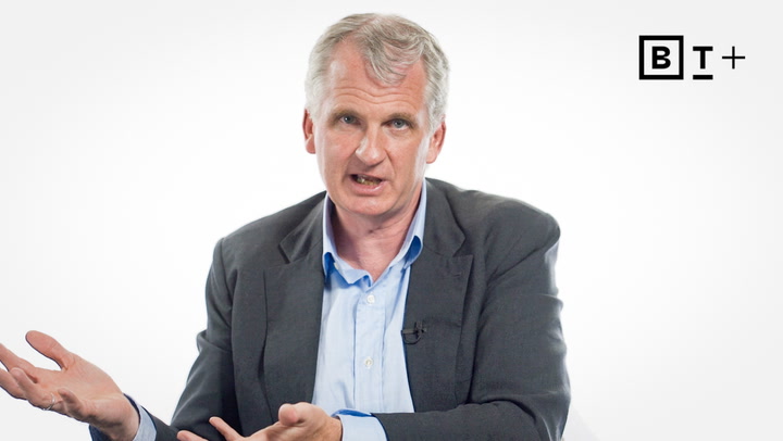 A middle-aged man in a suit jacket gestures with open hands while speaking, seated against a plain white background with the B T Plus logo in the top right corner.