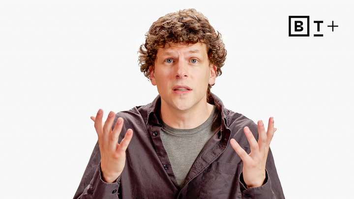 A person with curly hair gestures with both hands raised, wearing a dark shirt over a gray t-shirt, in front of a white background with a "B T +" logo in the top right corner.