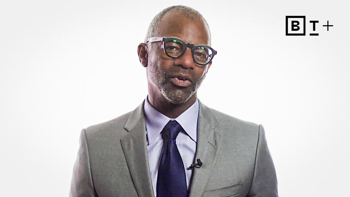 A man in a gray suit, blue shirt, and dark tie speaks in front of a white background with a "B T +" logo in the top right corner.