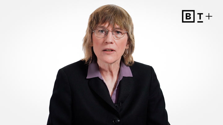 A person with shoulder-length light brown hair, glasses, and a black blazer over a purple shirt sits in front of a white background with a "B T +" logo in the top right corner.
