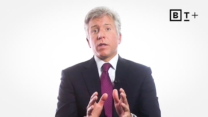 A man in a dark suit and purple tie speaks while gesturing with his hands, sitting against a plain white background with a "BT+" logo in the corner.