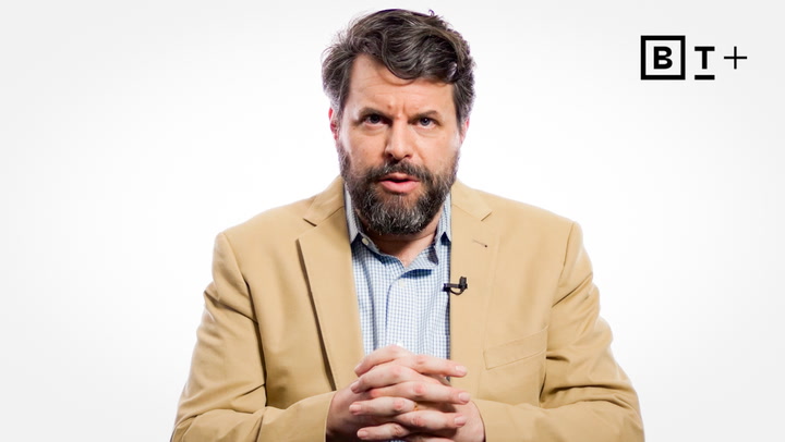 A man with a beard, wearing a tan blazer and blue shirt, sits with hands clasped against a white background. A black "B T +" logo appears in the upper right corner.