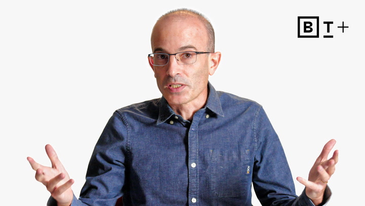 A middle-aged person with glasses and a denim shirt gestures with open hands while speaking, against a plain white background with a "B T +" logo in the corner.