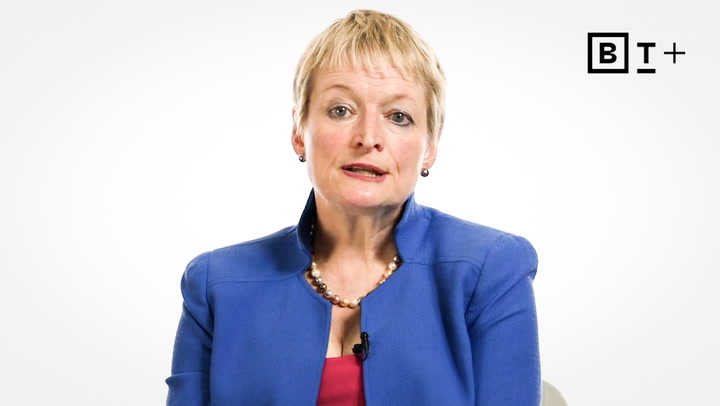 A woman with short blonde hair wearing a blue blazer, pink top, and pearl necklace sits against a white background with a "BT+" logo in the corner.