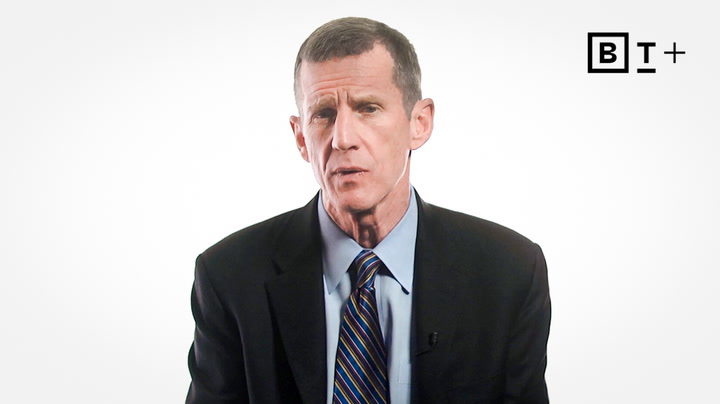 A man in a suit and striped tie speaks in front of a plain white background with a "B T +" logo in the top right corner.