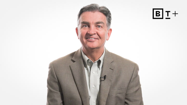 A man in a brown blazer and light shirt sits facing forward against a plain white background, with a BT+ logo in the upper right corner.