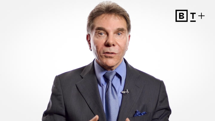 A man in a suit and tie speaks in front of a plain white background with a "BT+" logo in the top right corner.