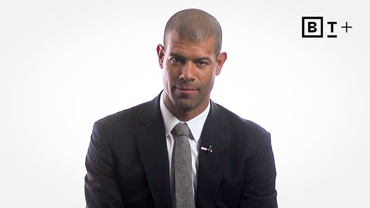 A man in a suit and tie sits against a plain white background with a "B T +" logo in the top right corner.