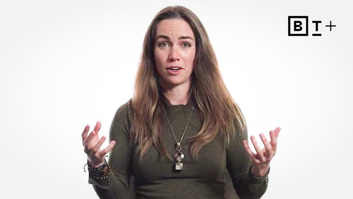 A woman with long brown hair wearing a green shirt and necklaces gestures with both hands. A "BT+" logo appears in the top right corner against a white background.