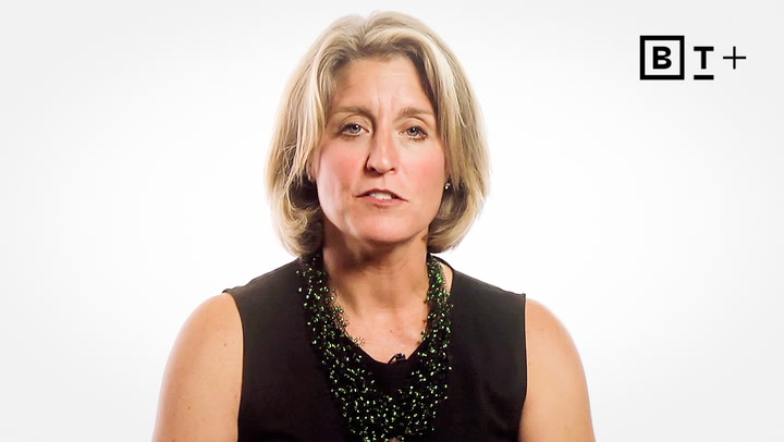 A woman with short blonde hair wearing a black sleeveless top and a green beaded necklace sits in front of a white background with a "BT+" logo in the top right corner.