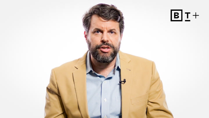A man with a beard and mustache wearing a tan blazer and blue shirt speaks in front of a white background with a "BT+" logo in the top right corner.