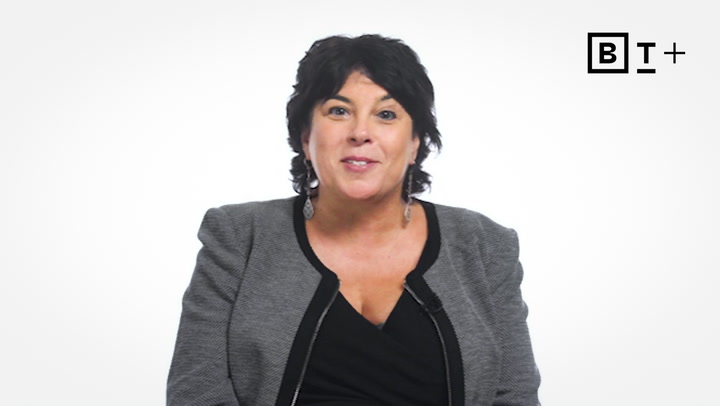 A woman with dark hair and a gray blazer smiles at the camera against a plain white background with a "B T +" logo in the top right corner.