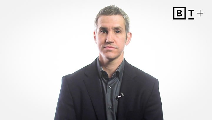 A man in a dark suit and collared shirt sits facing the camera against a white background with a BT+ logo in the top right corner.