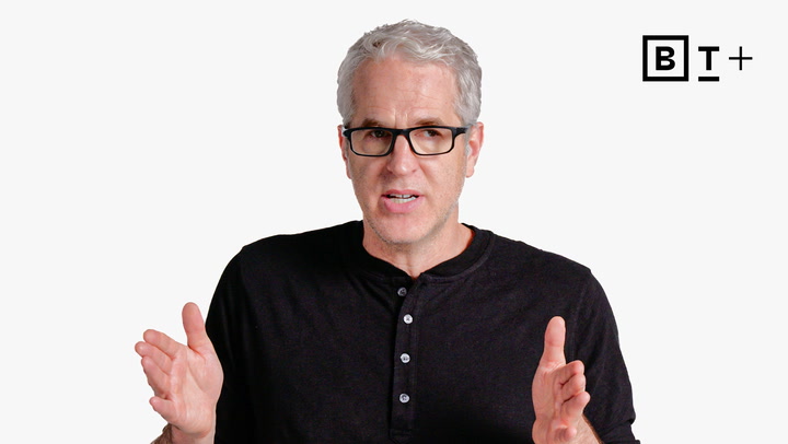 A man with gray hair and glasses, wearing a black henley shirt, gestures with his hands while speaking. A "B T +" logo appears in the top right corner.