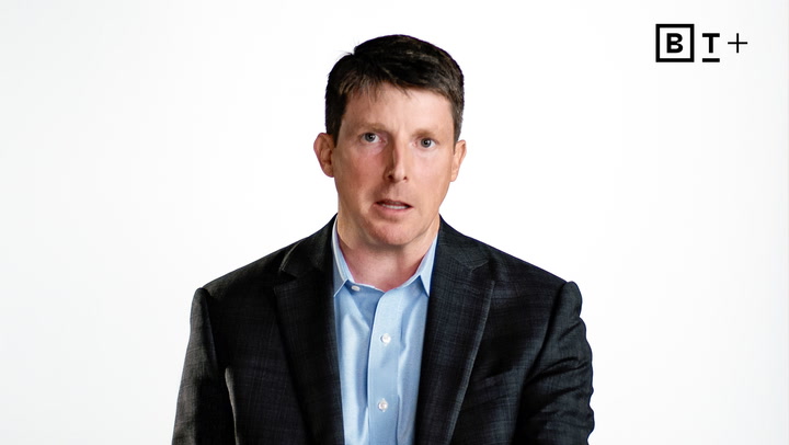 A man in a dark suit and light blue shirt sits against a plain white background with a "BT+" logo in the top right corner.