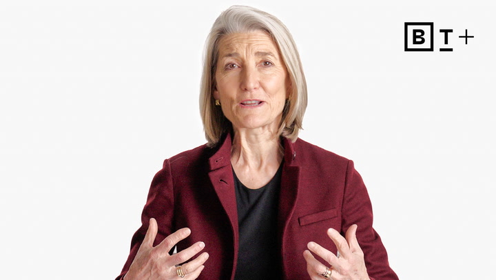 An older woman with gray hair, wearing a maroon blazer over a black shirt, gestures with her hands while speaking. A "B T +" logo appears in the top right corner.