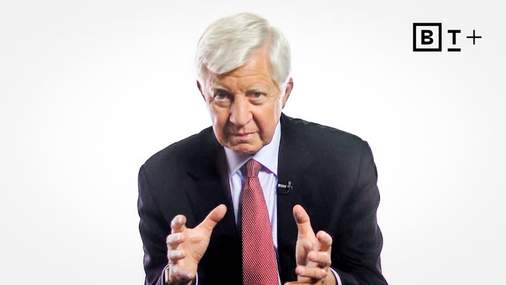 An older man in a suit and red tie speaks while gesturing with his hands; a "BT+" logo appears in the top right corner.