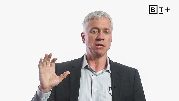A man in a suit speaks while raising his left hand, with a BT+ logo in the top right corner on a plain white background.