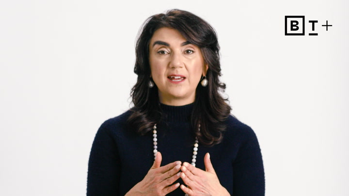 A woman with dark hair, wearing a dark sweater and a beaded necklace, speaks while gesturing with her hands; a "BT+" logo is in the top right corner.