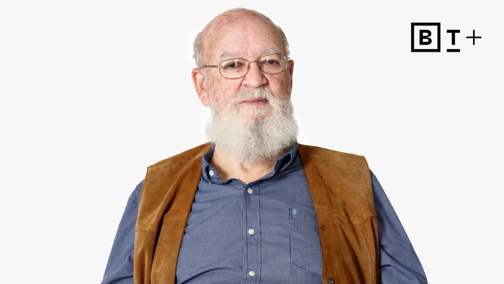 An older man with a white beard and glasses wears a blue shirt and brown vest, seated against a plain white background with a "BT+" logo in the top right corner.