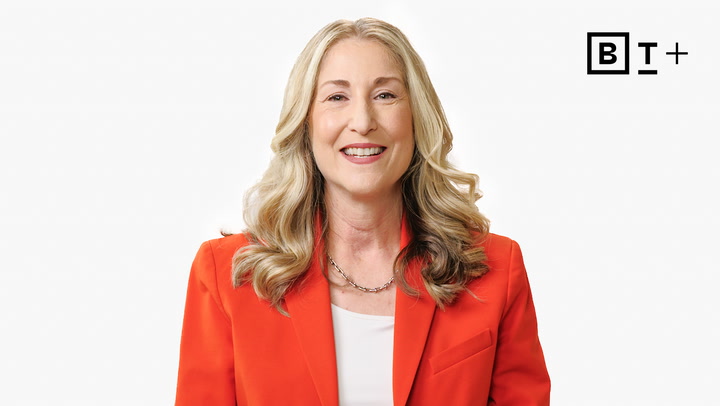 A woman with long blonde hair wearing an orange blazer smiles at the camera in front of a plain white background with a "BT+" logo in the top right corner.