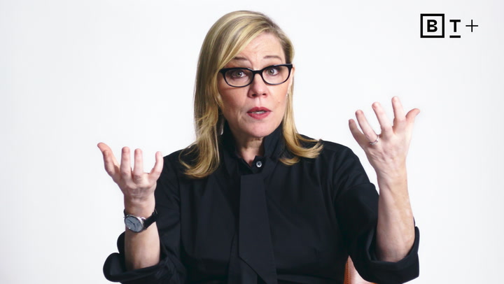 A woman with blonde hair and glasses sits against a white background, wearing a black top, and gesturing with both hands raised.