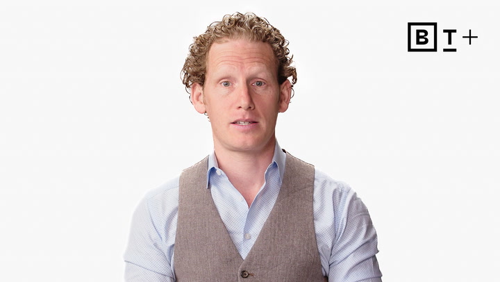 A man with curly hair, wearing a light blue shirt and brown vest, sits against a white background with the "B T +" logo in the top right corner.
