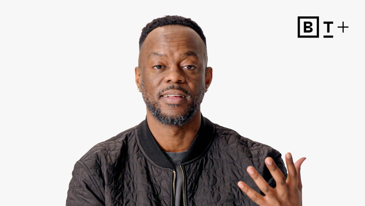 A man in a black quilted jacket gestures with his hand while speaking against a plain white background with a "B T +" logo in the top right corner.