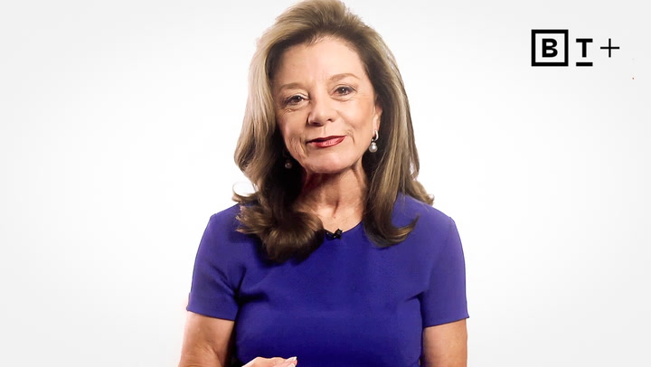 A woman with long brown hair wearing a purple dress stands in front of a white background with a “B T +” logo in the top right corner.